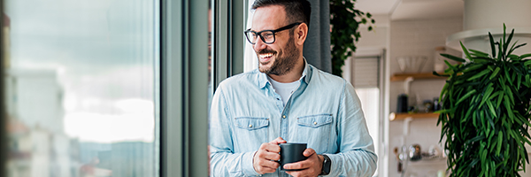 Smiling young adult businessman holding drinking coffee cup in office entrepreneur looking through window while standing in modern workplace man taking a time off or taking a brake from hard work