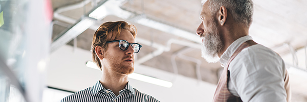 Low angle of concentrated male entrepreneurs in formal clothes looking at each other while standing in modern office with tablet during meeting about business plan together