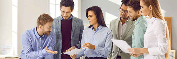 Team of diverse business people having a discussion at a work meeting. Group of colleagues standing by an office desk and listening to a young man explaining something in his report